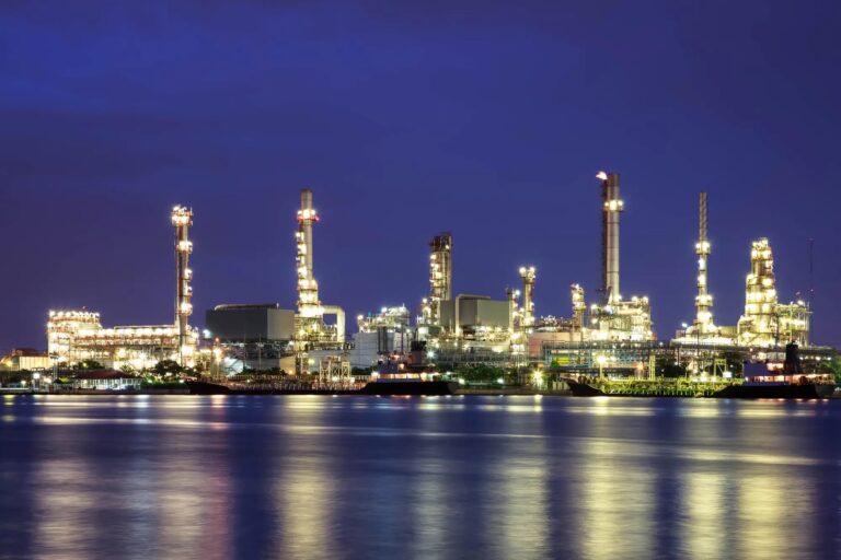 A petrochemical refinery lit up at twilight, reflected in the water in front of it